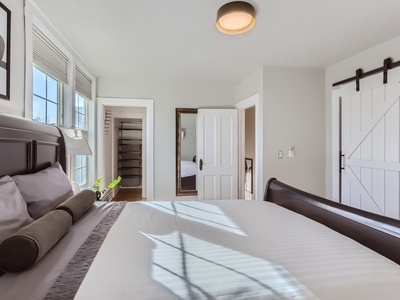 A bedroom with a neatly made bed, large windows, a mirror, and an open door leading to a closet. There is a barn door to the right and a ceiling light fixture.