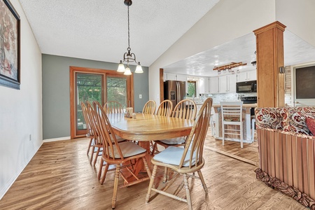 A dining area featuring a wooden table with eight chairs, adjacent to a kitchen with modern appliances, and an open living space with a partial view of a patterned sofa.