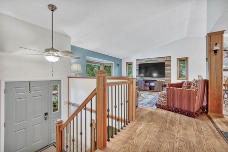 A split-level home entry features a staircase with wood railing leading to a living room with a striped sofa, a large TV on a wooden-paneled wall, and multiple windows. Ceiling fan above the entryway.