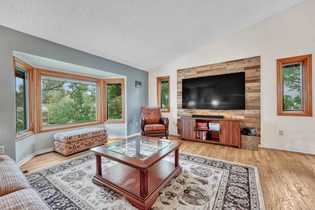 A living room with a large window, a wooden coffee table, a patterned rug, a wall-mounted TV on a wooden accent wall, and an armchair. The space is bright with natural light.