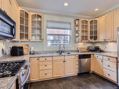 A modern kitchen with light wood cabinets, a granite countertop, and black tile floor. It features stainless steel appliances, a gas stove, microwave, dishwasher, toaster oven, and a window above the sink.