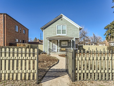 A small, light green, two-story house with white trim and a front porch, surrounded by a wooden picket fence, with a sidewalk leading to the front gate.