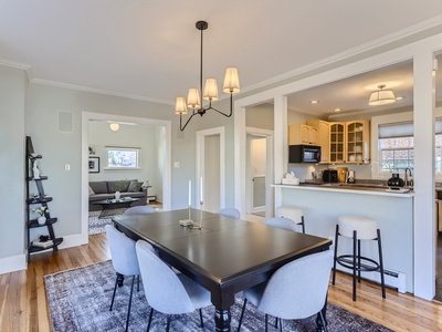 A modern dining room with a black table, six gray chairs, and a chandelier. To the right, there is a kitchen with a breakfast bar and stools. A living area is visible in the background.