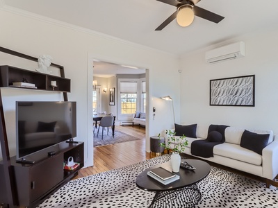 A modern living room features a white sofa with black cushions, a TV on a dark shelf, and a black ceiling fan. The room leads into a well-lit dining area. A patterned rug covers the floor.