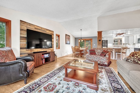 A spacious living area features a wall-mounted TV, patterned armchairs, a glass-top coffee table, a rug, and a view of the dining area and kitchen with white cabinets and wooden accents.