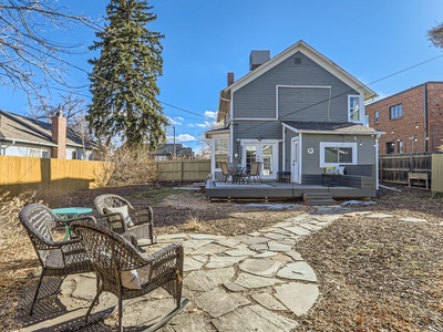 Backyard of a house with a stone pathway leading to a seating area with three wicker chairs and a table. The house features a patio with additional outdoor furniture and adjacent trees.