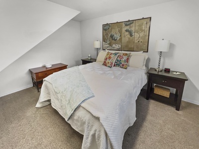 A bedroom with a white bedspread, patterned pillows, and a light blue blanket. The bed has two dark wood nightstands with lamps. A wooden chest is against the wall, and a decorative painting hangs above the bed.