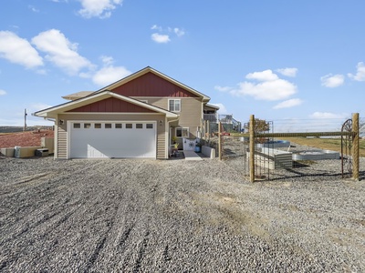 A house with a double garage and a gravel driveway sits under a blue sky. A fenced area with a gate is adjacent to the driveway.