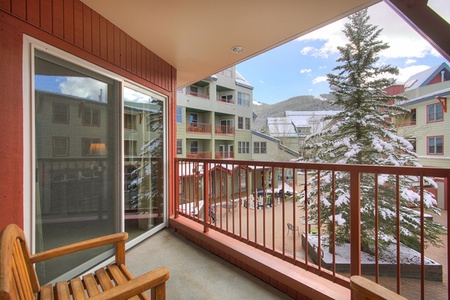 A balcony with a wooden bench overlooks a snow-covered courtyard with buildings, a pine tree, and mountain views in the distance under a partly cloudy sky.