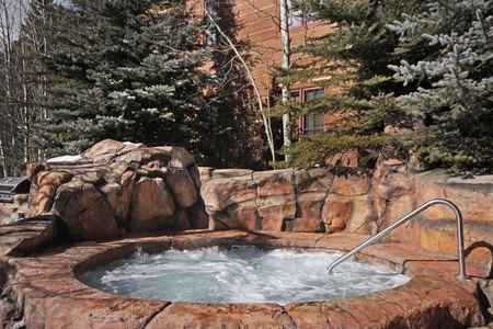 A stone hot tub with bubbling water is surrounded by evergreen trees and set against a rustic wooden building.