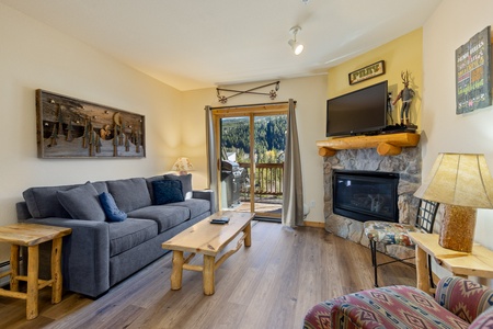 A cozy living room with a grey sofa, wooden furniture, a stone fireplace, and a mounted TV. Doors open to a balcony with a scenic forest view. Walls are adorned with rustic decor.