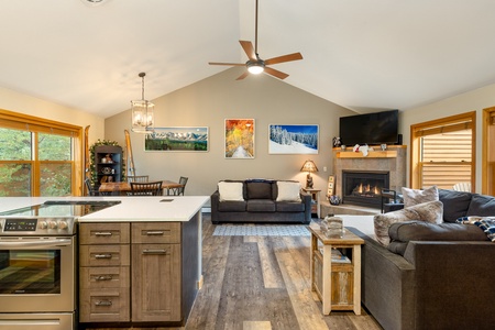 A modern open-concept living room and kitchen with wooden floors, a ceiling fan, photographs on the wall, a fireplace, beige and gray sofas, and a kitchen island with a stove and sink.