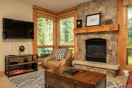 A cozy living room with a stone fireplace, wooden mantel, large windows, a leather armchair, a wooden coffee table, a TV mounted on the wall, and a shelf with decorative items.