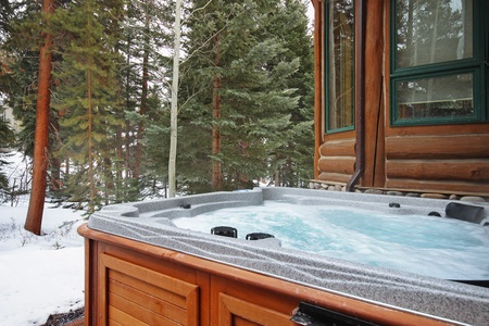 Outdoor hot tub with bubbling water next to a wooden cabin, surrounded by snow-covered ground and tall evergreen trees.