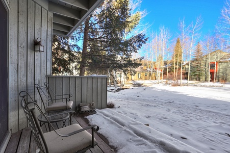 A wooden porch with two chairs overlooks a snowy backyard with trees and neighboring houses in the background on a clear day.