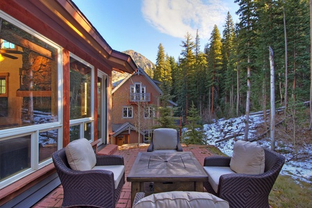 A patio with cushioned chairs and a table overlooks an alpine landscape with a classic wooden house, forested mountains, and patches of snow.