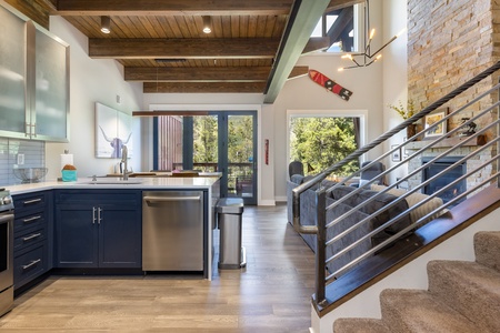 Modern kitchen with dark cabinets, stainless steel appliances, and a wooden beamed ceiling. Large windows and a staircase with a metal railing are visible.