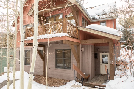 A two-story wooden house, with snow-covered roof and surroundings, features a balcony, intricate railing designs, and entrance with a few steps.