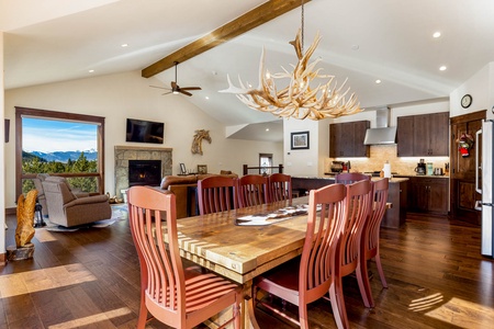 A spacious dining area with a wooden table, red chairs, an antler chandelier, adjacent to an open kitchen. A living area with a fireplace and large window overlooks a mountain landscape.