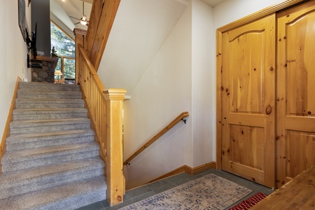 A hallway with a carpeted staircase leading upwards, wooden doors on the right, and a rug on a tiled floor in the foreground.