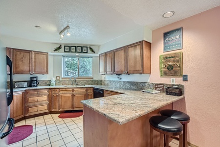 A kitchen with wooden cabinets, granite countertops, a black refrigerator, tiled floor, and two bar stools at a counter. Decor includes framed pictures and a small window over the sink.