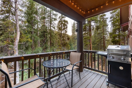 Covered wooden deck with two chairs, a round table, a grill, and string lights, overlooking a forest of tall pine trees.