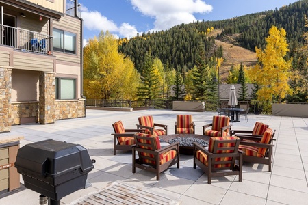 Outdoor seating area with a fire pit and a grill on a paved patio, surrounded by autumn trees and a hillside in the background.