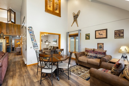 A cozy living and dining area featuring a round table with four chairs, two brown sofas, a wall-mounted snowboard, and various rustic decor and signs. A staircase leads to an upper loft.