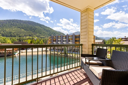 A balcony with two wicker chairs overlooks a serene lake and a backdrop of forested mountains on a sunny day.