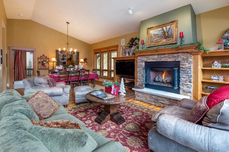 A cozy living room with cushioned sofas, a wooden coffee table, a stone fireplace, and a dining area in the background. The room is decorated with paintings, candles, and holiday-themed items.