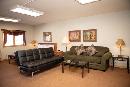 A living room area with a black futon, a green sofa, two coffee tables, two lamps, and framed artwork on a beige wall. A bed is visible in the background near a window with red curtains.