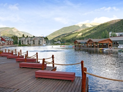 Lakeside wooden deck with benches overlooking a calm lake and mountainous backdrop. Buildings and docks with boats are seen along the shoreline.