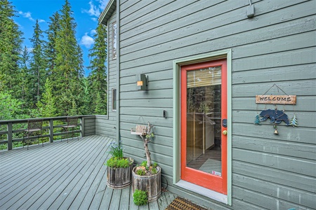 Green wooden house with a red door, two potted plants, and a "Welcome" sign featuring a black bear. A wooden deck with railings surrounds the house, overlooking a forested area.