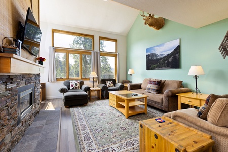 A cozy living room with large windows, two sofas, two armchairs, a coffee table, a rug, a fireplace, a mounted deer head, and a mountain landscape painting on a green accent wall.