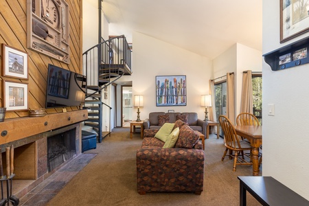 A cozy living room with a spiral staircase, fireplace, couch, chairs, and a dining table. The room features a high ceiling and large windows with curtains.