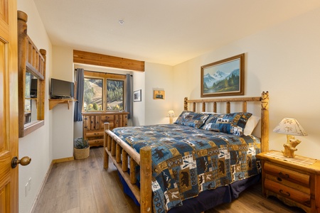 A cozy bedroom features a wooden bed with a patchwork quilt, wooden furniture, a small TV, a window with mountain views, and a painting above the bed.