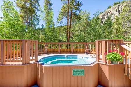 Outdoor hot tub surrounded by a wooden fence and greenery, located in a mountainous area. A small sign reads "No Glass Allowed In Pool Area." Potted plants with red flowers adorn the fence.