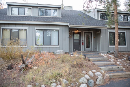 A two-story gray house with a porch, several windows, and a sloped roof, surrounded by rocks and trees, with a small staircase leading to the front door.