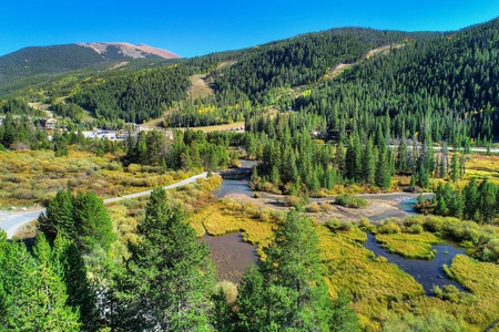 A scenic landscape featuring a river winding through a lush, green valley with forested mountains in the background under a clear blue sky. A road runs along the valley floor.