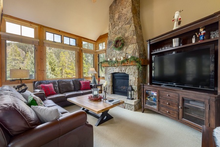 A cozy living room with a large stone fireplace, leather sofas, a decorated Christmas mantel, a large TV, and large windows offering views of trees outside.