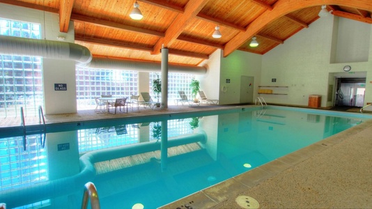 Indoor swimming pool area with natural light from large windows. Features deck chairs, tables, and potted plants. The ceiling has wooden beams and there is a door leading to another room.