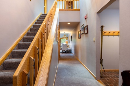 A carpeted stairway with wooden railings leads to an upper level. Next to it, a hallway with framed pictures on the wall leads to a brightly lit room with a window. A coat rack stands at the entrance.