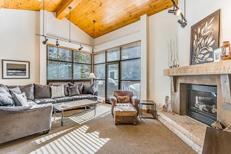 A cozy living room with a large sectional sofa, armchair, fireplace, coffee table, and tall windows with blinds, featuring a high wooden ceiling and minimalistic decor.