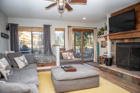 A cozy living room features a sectional sofa with cushions, a large ottoman, a fireplace with a TV above it, windows with curtains, and a sliding glass door leading to an outdoor area.