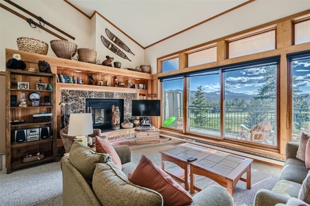 A cozy living room features a stone fireplace, a mounted TV, large windows offering a scenic outdoor view, and wooden shelves filled with various decorative items and woven baskets.