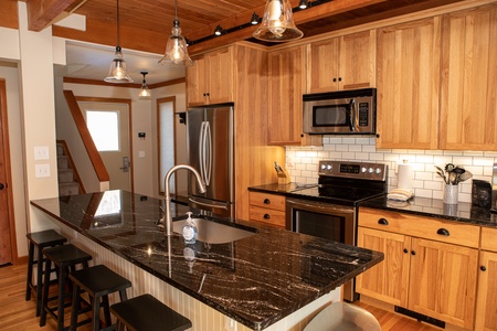 A modern kitchen with wooden cabinets, a black granite island countertop, stainless steel appliances, and pendant lighting. The space includes a sink, oven, microwave, and stools.