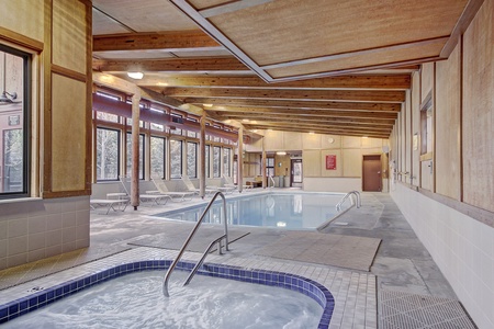 Indoor pool area with large windows, a hot tub in the foreground, a pool in the background, and wooden ceiling beams. Several lounge chairs are against the windows.