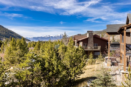 Houses with wooden exteriors are nestled among trees with mountains in the background under a blue sky.