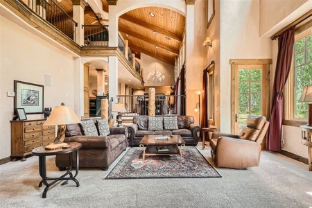 Spacious living room with high wooden ceilings, large windows, and a mix of leather and fabric furniture. It features a loft area, chandelier, and patterned rug on a carpeted floor.