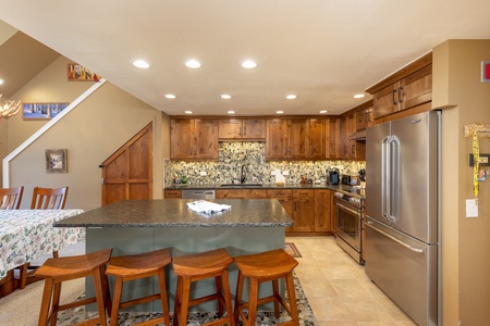 A modern kitchen with wooden cabinets, stainless steel appliances, a large island with a granite countertop, and four wooden bar stools. Recessed lighting illuminates the space.